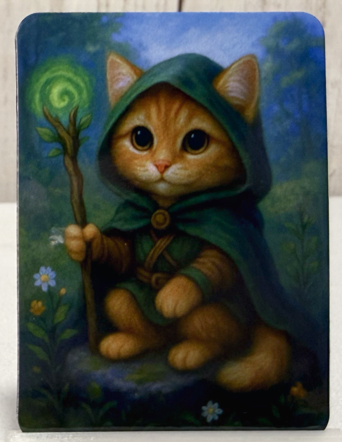 Magnet featuring a druid cat in a green hood holding a staff with a spiral design, surrounded by nature.