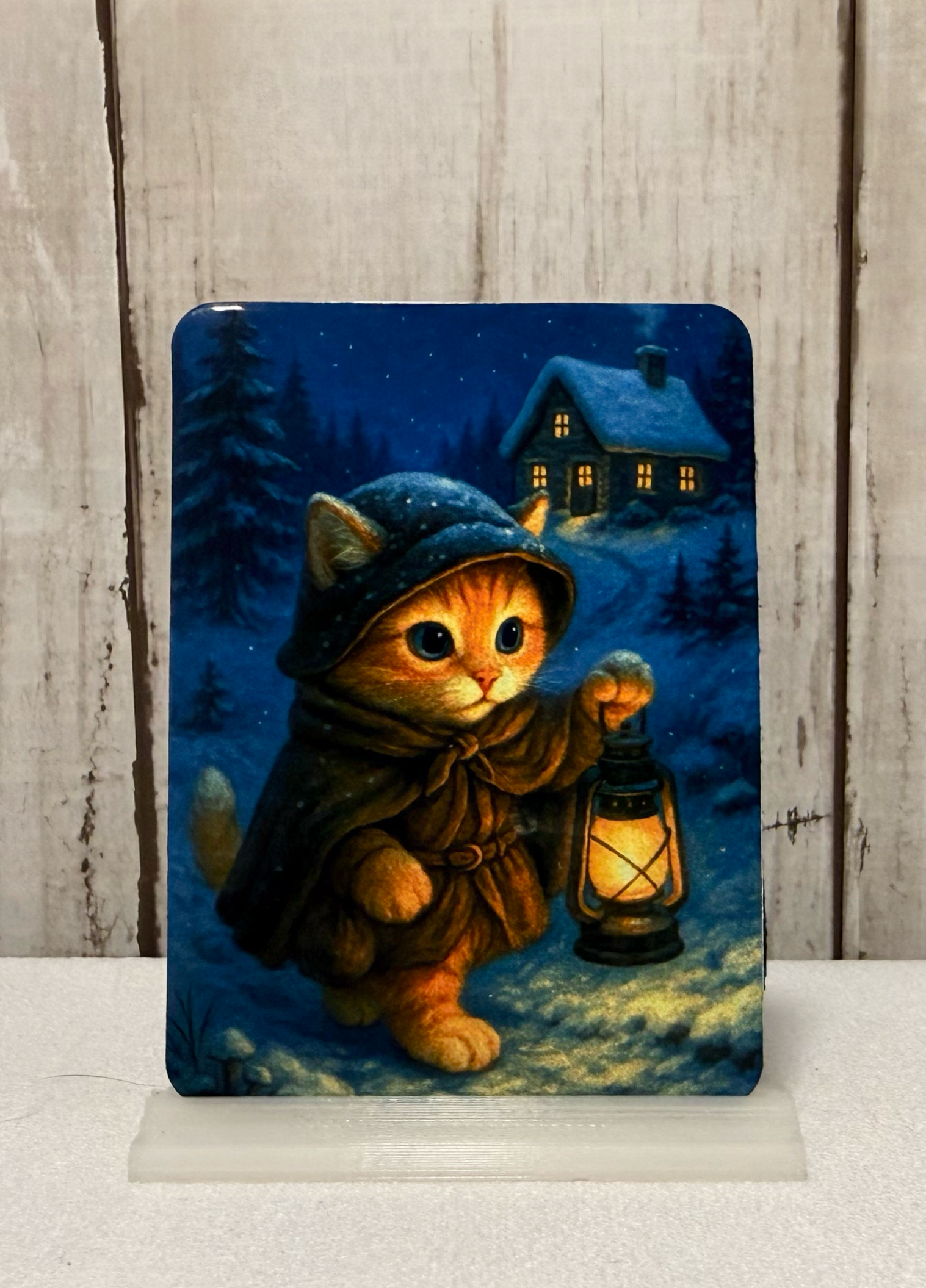 Magnet featuring a cat in a hood and coat holding a lantern in a snowy landscape with a house in the background.