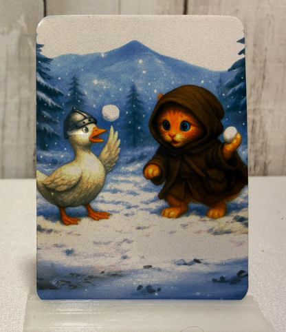 Magnet featuring a cat in a hooded outfit and a goose playing with snowballs in a snowy landscape.